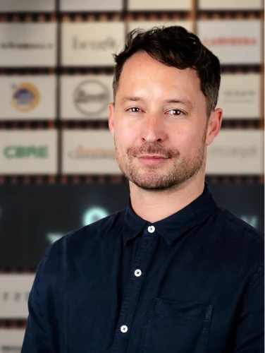 Simon, Chief Customer Officer at Online Marketing Gurus, in a navy button-up shirt in front of the OMG client logo wall.