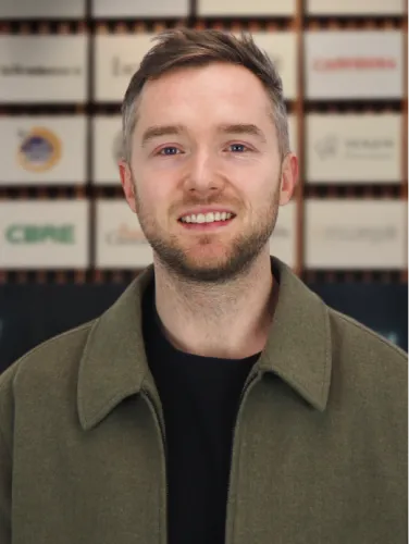 The Paid Social Director at Online Marketing Gurus, smiling in an olive green jacket in front of the OMG client logo wall.