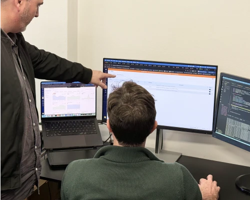 Two Online Marketing Gurus developers collaborating at a multi-monitor workstation, reviewing code and a web project across laptop and external displays.
