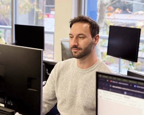 An Online Marketing Gurus team member working at a multi-monitor desk by a window, managing website hosting infrastructure.